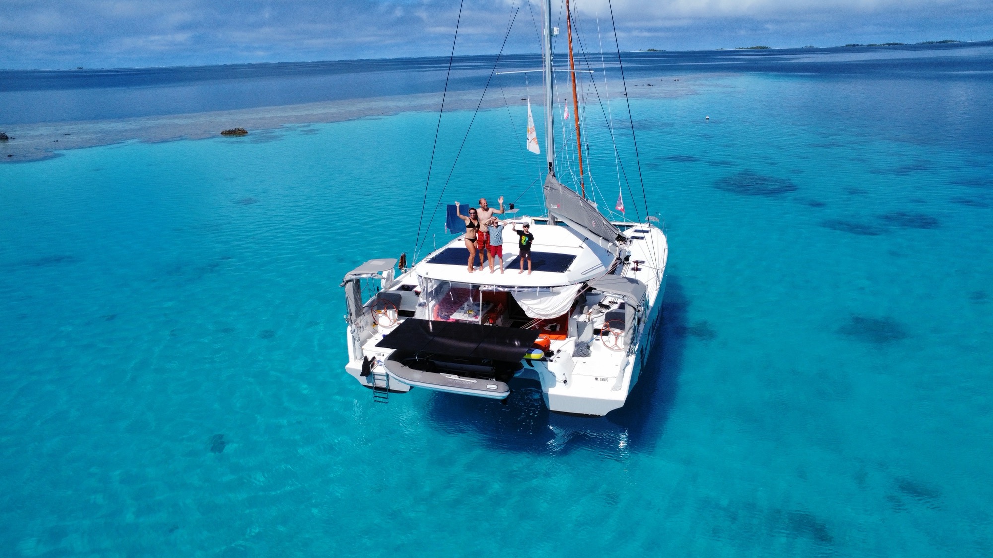 Audrey, Nicolas, Noah et Léo à bord de Pélican, saluant depuis l'étrave, lagon turquoise de Polynésie française